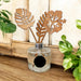 A reed diffuser bottle with a clear glass base, burlap wrap, and two wooden leaf reeds, styled on a wooden surface with green plants and a rustic paneled background.