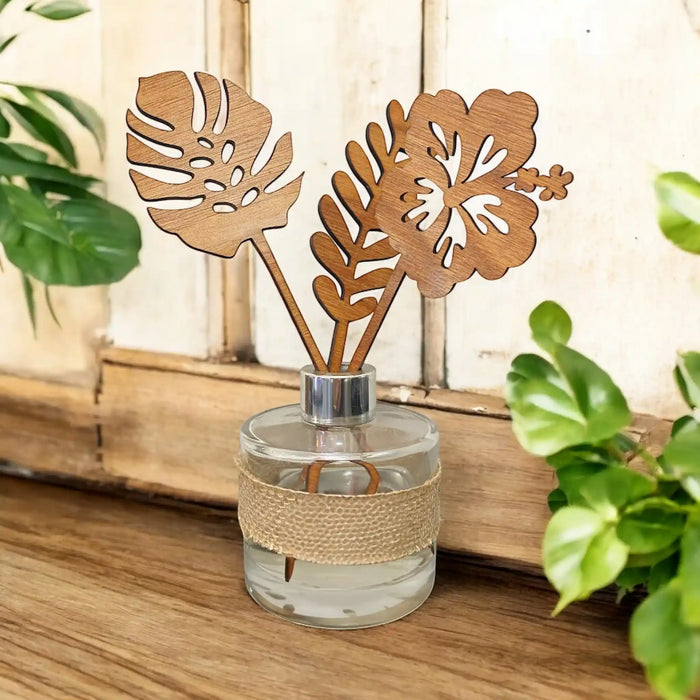 A glass reed diffuser bottle with a beige jute wrap and two laser-cut wooden reeds shaped like tropical leaves, styled on a wooden surface with green plants in the background.