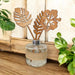 A glass reed diffuser bottle with a beige jute wrap and two laser-cut wooden reeds shaped like tropical leaves, styled on a wooden surface with green plants in the background.