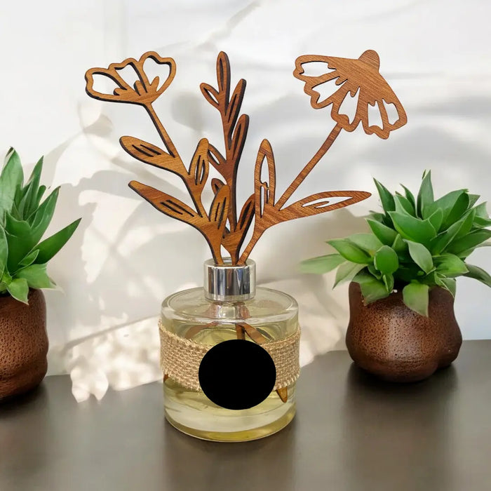 A glass reed diffuser bottle with a silver cap and a decorative wooden flower stem reed, styled on a dark tabletop with two potted succulents in the background.