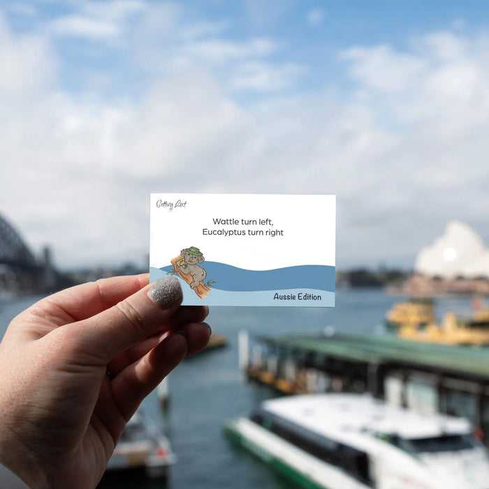 Hand holding a card with a cartoon character and text, blurred background of a harbor with boats and a bridge.