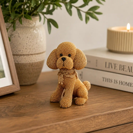 A small poodle-shaped decorative figurine made of gold-toned beads sits on a wooden tabletop, styled with a vase of greenery, books, and a lit candle in the background.
