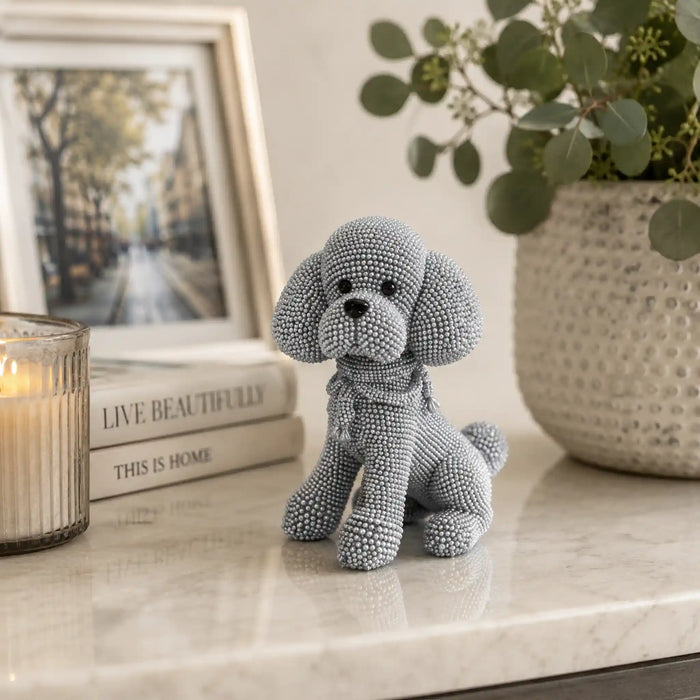 A small gray beaded poodle figurine sits on a light marble surface in a styled interior setting, with a candle, books, framed picture, and potted plant in the background.