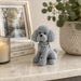 A small gray beaded poodle figurine sits on a light marble surface in a styled interior setting, with a candle, books, framed picture, and potted plant in the background.