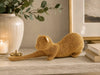 A gold beaded cat figurine posed stretching on a light marble surface, styled in a warm home interior with a white vase, candle, framed art, and books in the background.