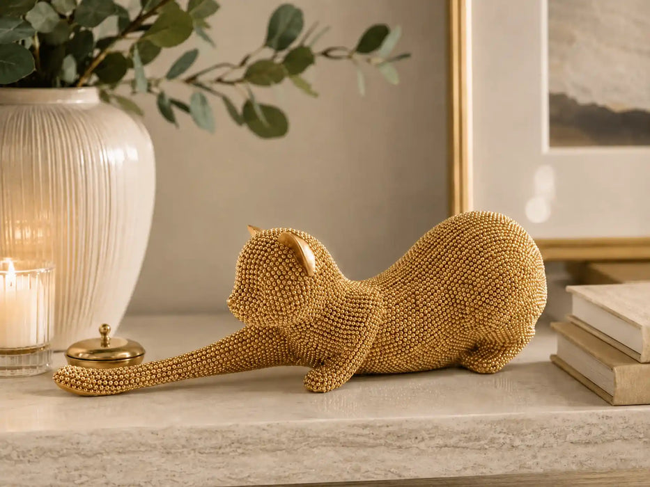 A gold beaded cat figurine posed stretching on a light marble surface, styled in a warm home interior with a white vase, candle, framed art, and books in the background.