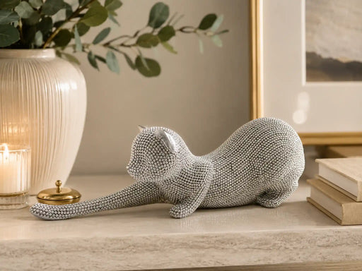 A sculptural cat figurine covered in small silver rhinestones, posed stretching on a light stone mantel with a cream vase, candle, and framed art in the background.
