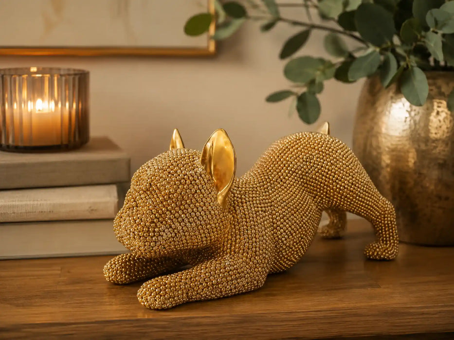A decorative figurine of a French Bulldog lying down, covered in small gold beads with polished gold ears, displayed on a wooden table in a warm, styled interior setting.
