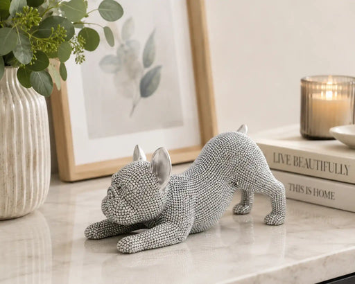 A textured gray French Bulldog figurine in a crouching pose, styled on a marble tabletop with a vase of greenery, a framed print, and a candle in the background.