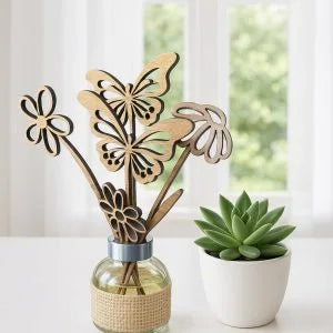 Laser-cut wooden butterfly and flower diffuser reeds in a glass bottle with a beige woven wrap, shown next to a small potted succulent in a bright room.