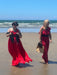 Two women walking on a sandy beach with the ocean and blue sky in the background. The woman on the left wears a flowing bright red dress and a dark hat, while the woman on the right wears a matching red jumpsuit with a black belt and carries a tan tote bag.