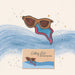 A stylized illustrated product shot showing a small rectangular box labeled 'Getting Lost Girls Road Trip Edition' with matching sunglasses above it, set against a cream background with blue watercolor waves and small gold star accents.