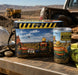 A construction-themed insulated cooler bag and matching can cooler sit on a work surface in a desert construction site. The products feature a printed scene of construction workers sitting in chairs facing a desert landscape, with yellow warning-style graphics and the Screw It brand visible on both items.