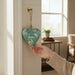 Hand holding a teal floral metal heart decoration hanging on a door hook in a warm home interior