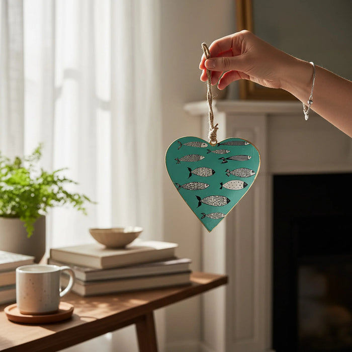 A hand holds a heart-shaped teal ornament decorated with silver fish silhouettes, shown in a softly lit living room setting with books, a mug, and a plant on a table.