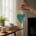 A hand holds a heart-shaped teal ornament decorated with silver fish silhouettes, shown in a softly lit living room setting with books, a mug, and a plant on a table.