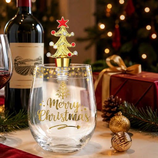 A clear stemless wine glass with gold 'Merry Christmas' lettering and a gold tree design, topped with a gold Christmas tree-shaped bottle stopper featuring a red star. The scene is styled with a wine bottle, glass, wrapped gift, pinecones, ornaments, and a blurred Christmas tree in the background.