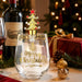 A clear stemless wine glass with gold 'Merry Christmas' lettering and a gold tree design, topped with a gold Christmas tree-shaped bottle stopper featuring a red star. The scene is styled with a wine bottle, glass, wrapped gift, pinecones, ornaments, and a blurred Christmas tree in the background.