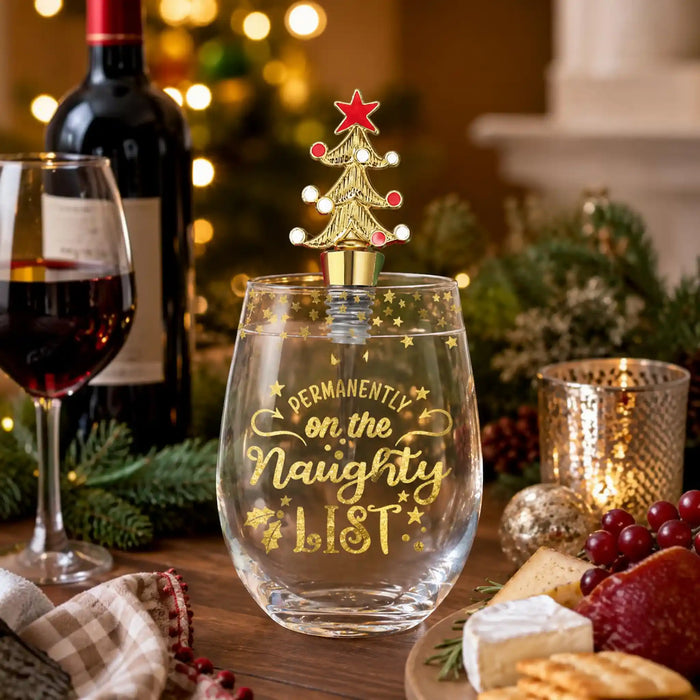 A clear stemless wine glass with gold lettering reading 'PERMANENTLY on the Naughty LIST' and a gold Christmas tree bottle stopper with a red star, shown in a festive holiday setting with wine, cheese, crackers, and warm lights.
