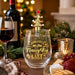 A clear stemless wine glass with gold lettering reading 'PERMANENTLY on the Naughty LIST' and a gold Christmas tree bottle stopper with a red star, shown in a festive holiday setting with wine, cheese, crackers, and warm lights.