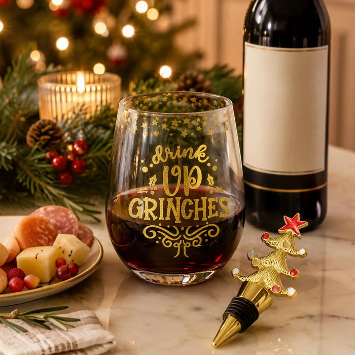 A festive holiday wine glass with gold lettering that reads 'drink UP GRINCHES' filled with red wine, styled on a marble table with a wine bottle, a Christmas tree-shaped wine stopper, and holiday decorations in the background.