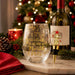 A clear stemless wine glass with gold holiday text reading 'It's 5 o'clock at the North Pole' is styled on a marble surface next to a wine bottle and a gold Christmas tree wine stopper, with a blurred Christmas tree and holiday decorations in the background.