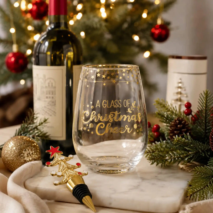 A clear stemless wine glass with gold lettering that reads 'A GLASS OF Christmas Cheer' is styled on a marble surface with a wine bottle, Christmas tree lights, pine branches, red berries, and a gold bottle stopper in a festive holiday setting.