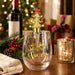 A clear stemless wine glass with gold holiday lettering reading 'Dear Santa, Have Some Wine' and a gold Santa tree dispenser with a red star topper resting in it, set on a wooden table with Christmas decorations, a wine bottle, and wrapped gifts in the background.