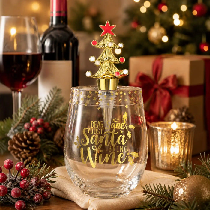 A clear stemless wine glass with gold lettering that reads 'Dear Jane, Just Santa Bring Wine,' topped with a gold Christmas tree and red star. The glass is styled on a table with holiday decor, a wine bottle, a glass of red wine, candles, pinecones, and wrapped gifts in the background.