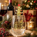 A clear stemless wine glass with gold lettering that reads 'Dear Jane, Just Santa Bring Wine,' topped with a gold Christmas tree and red star. The glass is styled on a table with holiday decor, a wine bottle, a glass of red wine, candles, pinecones, and wrapped gifts in the background.