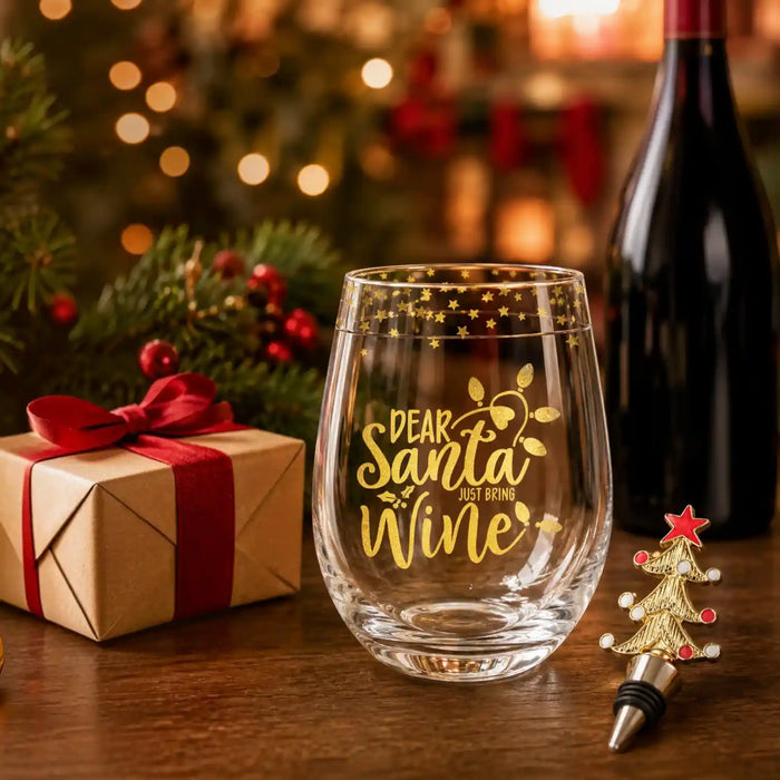 A clear stemless wine glass with gold 'Dear Santa Just Bring Wine' lettering and gold star details, styled on a wooden table with a wrapped gift, a wine bottle, and a small Christmas tree bottle stopper in a festive holiday setting.