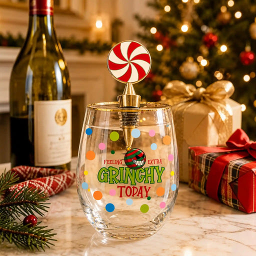 A clear stemless wine glass with colorful polka dots and the phrase 'FEELING EXTRA GRINCHY TODAY' in green and red, topped with a gold and silver stopper shaped like a candy cane. The glass is set on a marble surface with a blurred Christmas tree, wrapped gifts, a wine bottle, and pine branches in the background.