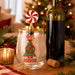 A clear stemless wine glass with a festive Christmas design, featuring a green tree under a red prohibition sign and the words '100% that PERSON!'. The glass is topped with a gold stopper shaped like a red-and-white peppermint candy.