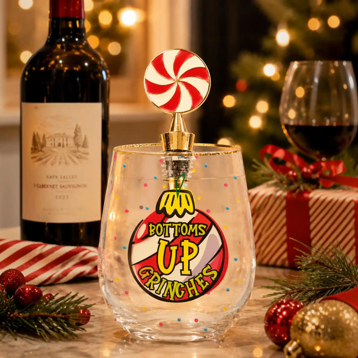 A clear stemless wine glass with festive multicolor confetti and a large Christmas ornament graphic reading 'BOTTOMS UP CRUNCHES,' topped with a candy cane topper. The glass is styled on a tabletop with a wine bottle, wine glass, wrapped gift, ornaments, and blurred Christmas lights in the background.