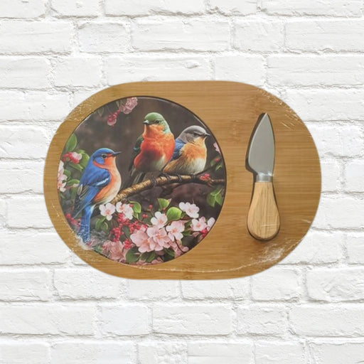 An oval wooden serving board with a printed circular design of colorful birds perched among pink flowers, shown on a white brick wall next to a small knife with a wooden handle.