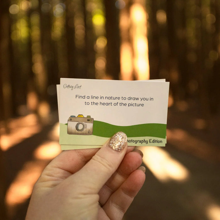 A hand with glittery nail polish holds a small printed card in a sunlit forest. The card reads, 'Find a line in nature to draw you in to the heart of the picture' with a small camera illustration and the text 'photography Edition' near the bottom.