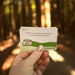 A hand with glittery nail polish holds a small printed card in a sunlit forest. The card reads, 'Find a line in nature to draw you in to the heart of the picture' with a small camera illustration and the text 'photography Edition' near the bottom.