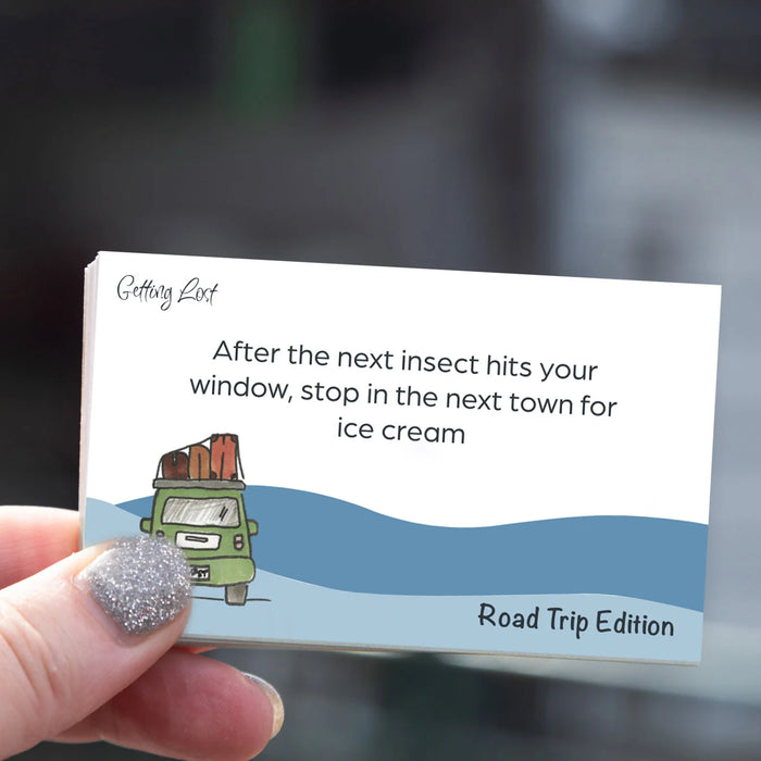 Hand holding a card with a humorous road trip message and illustration of a car with luggage.