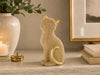 A gold-toned cat figurine covered in small clear rhinestones, sitting on a marble shelf next to a lit candle, a white vase with greenery, and stacked books.