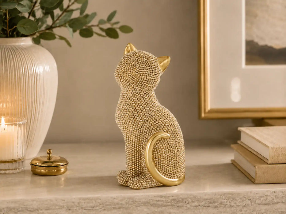 A gold-toned cat figurine covered in small clear rhinestones, sitting on a marble shelf next to a lit candle, a white vase with greenery, and stacked books.