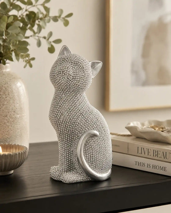 A silver cat figurine covered in small rhinestones, sitting on a dark wooden table next to a candle, a vase with greenery, and a stack of books.