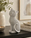 A silver cat figurine covered in small rhinestones, sitting on a dark wooden table next to a candle, a vase with greenery, and a stack of books.