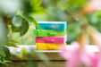 Four small rectangular storage boxes stacked on a wooden surface, shown outdoors with blurred green leaves and pink flowers in the foreground. The boxes are bright blue, green, pink, and yellow.