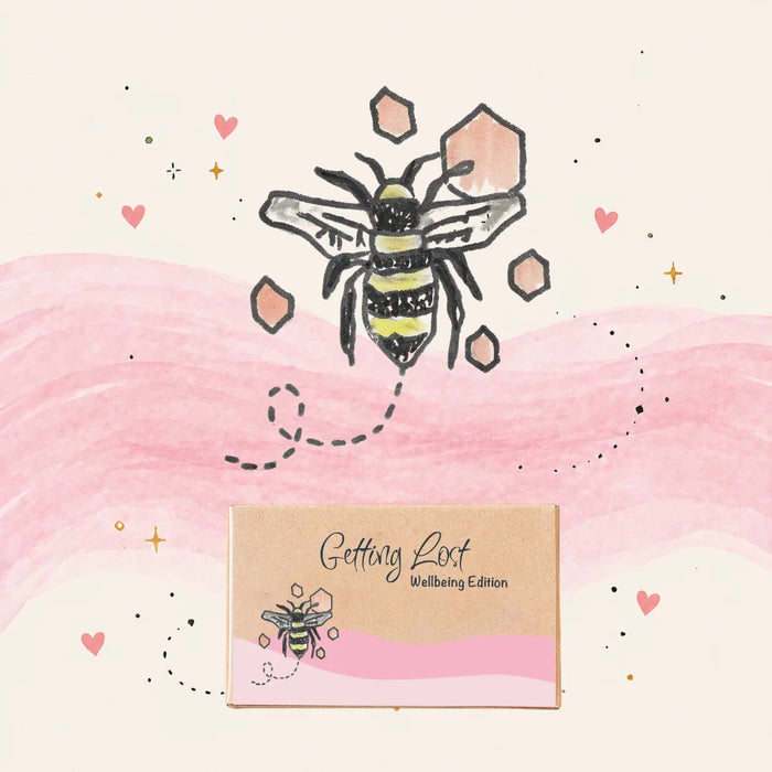 A kraft paper gift box labeled 'Getting Lost Wellbeing Edition' with a pink watercolor-style band and bee illustration, shown on a cream background with a larger bee drawing, pink hills, and small gold stars.