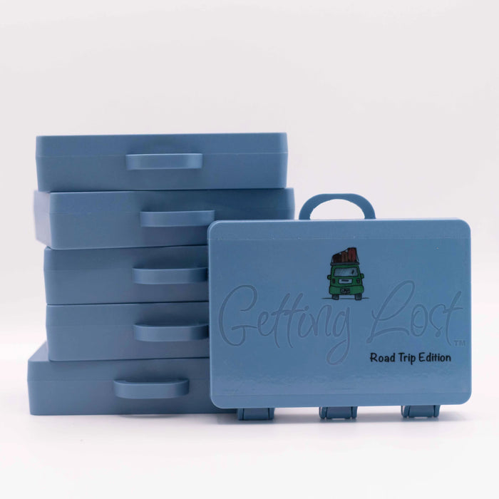 A stack of five blue plastic storage boxes with handles sits next to one open-front box in the foreground. The front box has a handle, embossed script reading 'Getting Lost,' and small text reading 'Road Trip Edition' with a small green truck graphic.