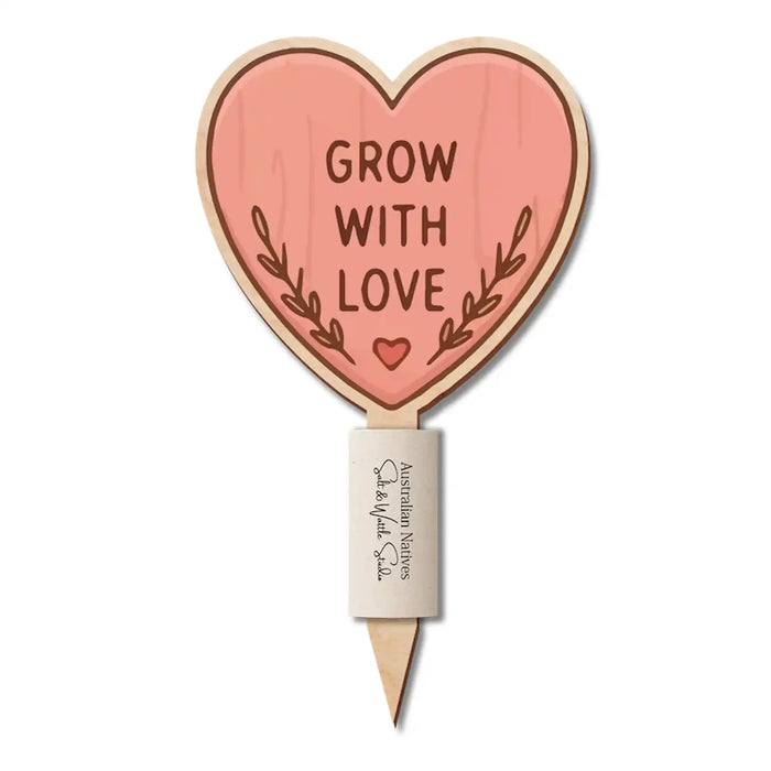 A heart-shaped wooden garden stake with a pinkish-orange face that reads 'GROW WITH LOVE' in brown lettering, surrounded by a small leaf motif. The stake has a natural wood grain border and a white label near the pointed bottom.