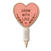 A heart-shaped wooden garden stake with a pinkish-orange face that reads 'GROW WITH LOVE' in brown lettering, surrounded by a small leaf motif. The stake has a natural wood grain border and a white label near the pointed bottom.