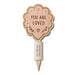 A wooden 'You Are Loved' cake topper with a scalloped leaf-shaped top featuring engraved text and a small heart, mounted on a pointed stick with a white paper sleeve printed with 'Aurora Villes Cake Toppers'.