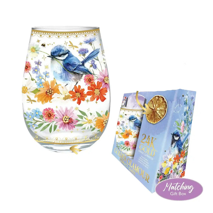 A stemless wine glass and matching gift box featuring a bluebird and colorful wildflower illustration. The glass has gold decorative trim, and the box is light blue with gold text and a gold dried citrus slice accent.