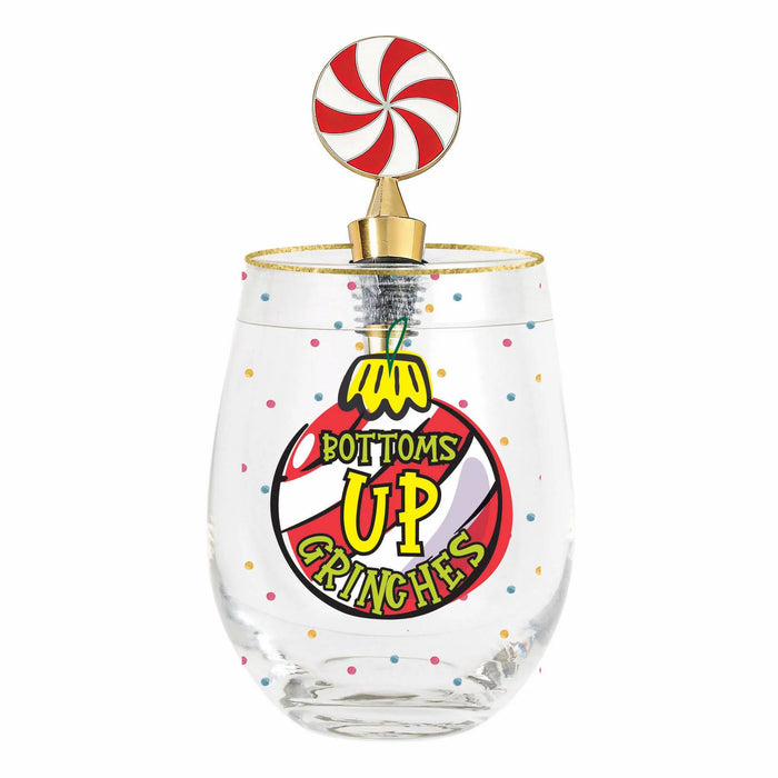 A clear stemless wine glass with a gold rim, decorated with colorful confetti dots and a cartoon ornament graphic that reads 'BOTTOMS UP CRINCHES' in yellow and red. The glass is topped with a gold-colored metal stand holding a red and white swirled candy cane ornament.